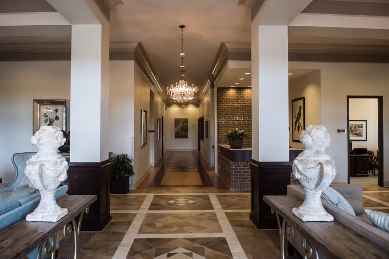 Interior view of a senior living facility hallway with a chandelier hanging from the ceiling. The hallway is flanked by columns and decorated with framed artwork on the walls. There are two white decorative urns on wooden tables in the foreground, and seating areas with sofas are partially visible on both sides.