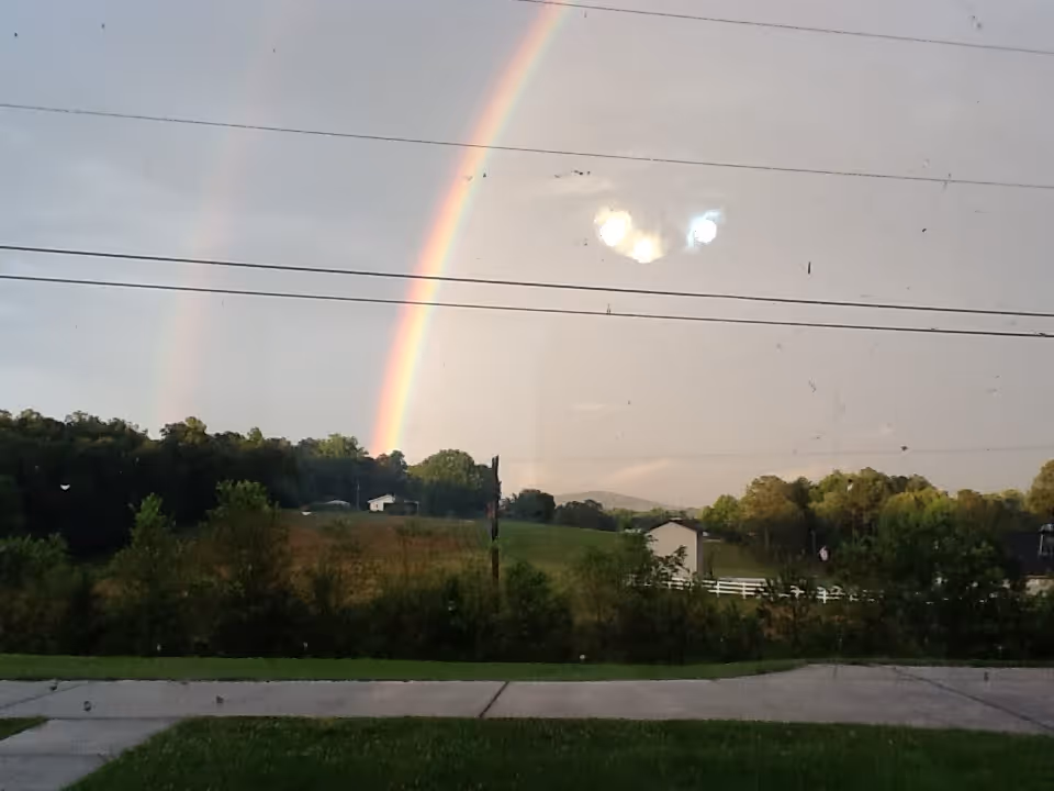 A double rainbow arches over a rural field with trees and power lines, seen through a window.