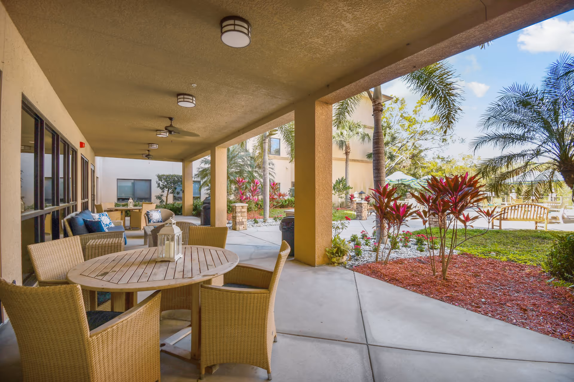 Covered outdoor patio area with round wooden tables and wicker chairs. The patio overlooks a landscaped garden with palm trees, red-leafed plants, and a bench. The sky is blue with some clouds.