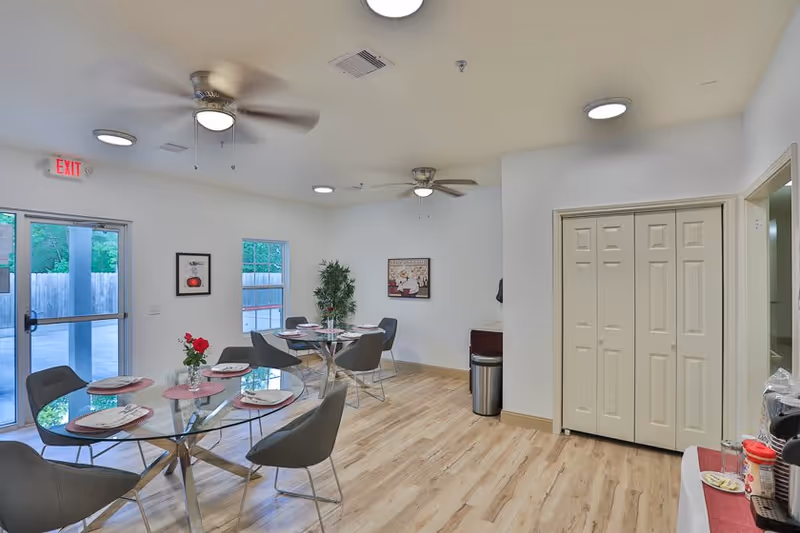 A bright dining area with two glass tables set with plates and napkins, surrounded by gray chairs. The room has light wood flooring, white walls, ceiling fans, and recessed lighting. There are two windows and a glass door leading outside, a potted plant in the corner, and a small counter with a trash can and some items on the right side.
