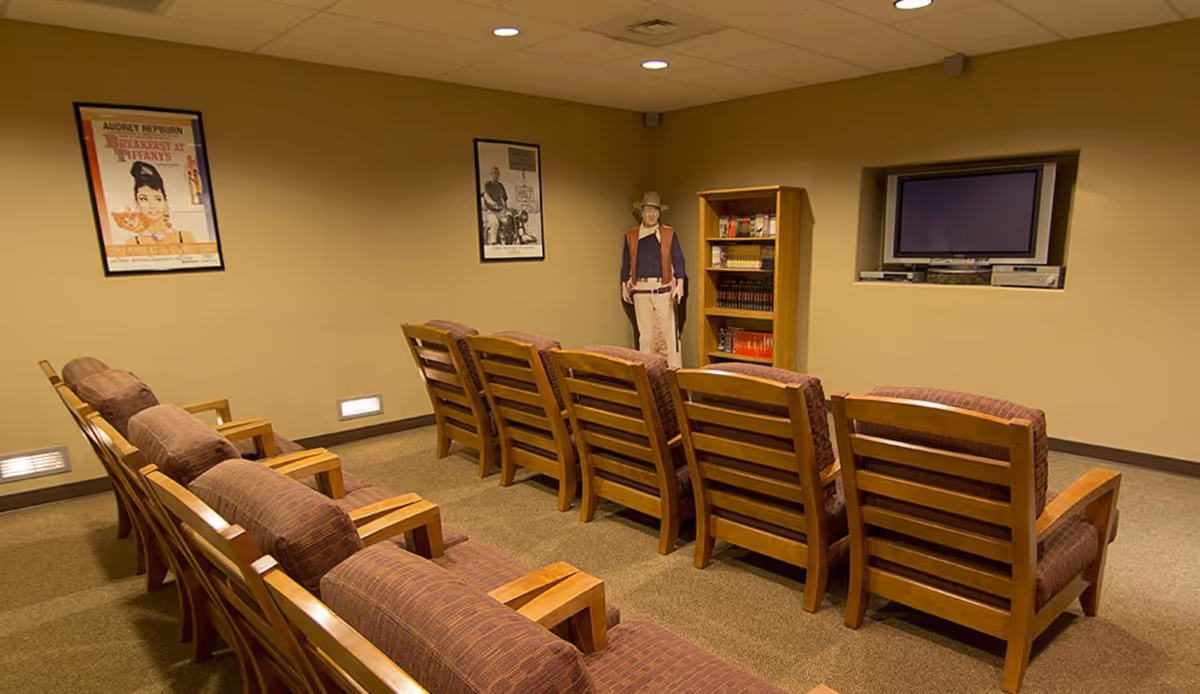 A small theater room with two rows of wooden chairs with cushioned seats facing a wall-mounted flat screen TV. The room has beige walls with two framed movie posters, a bookshelf filled with books, and a life-size cardboard cutout of a cowboy figure in the corner.