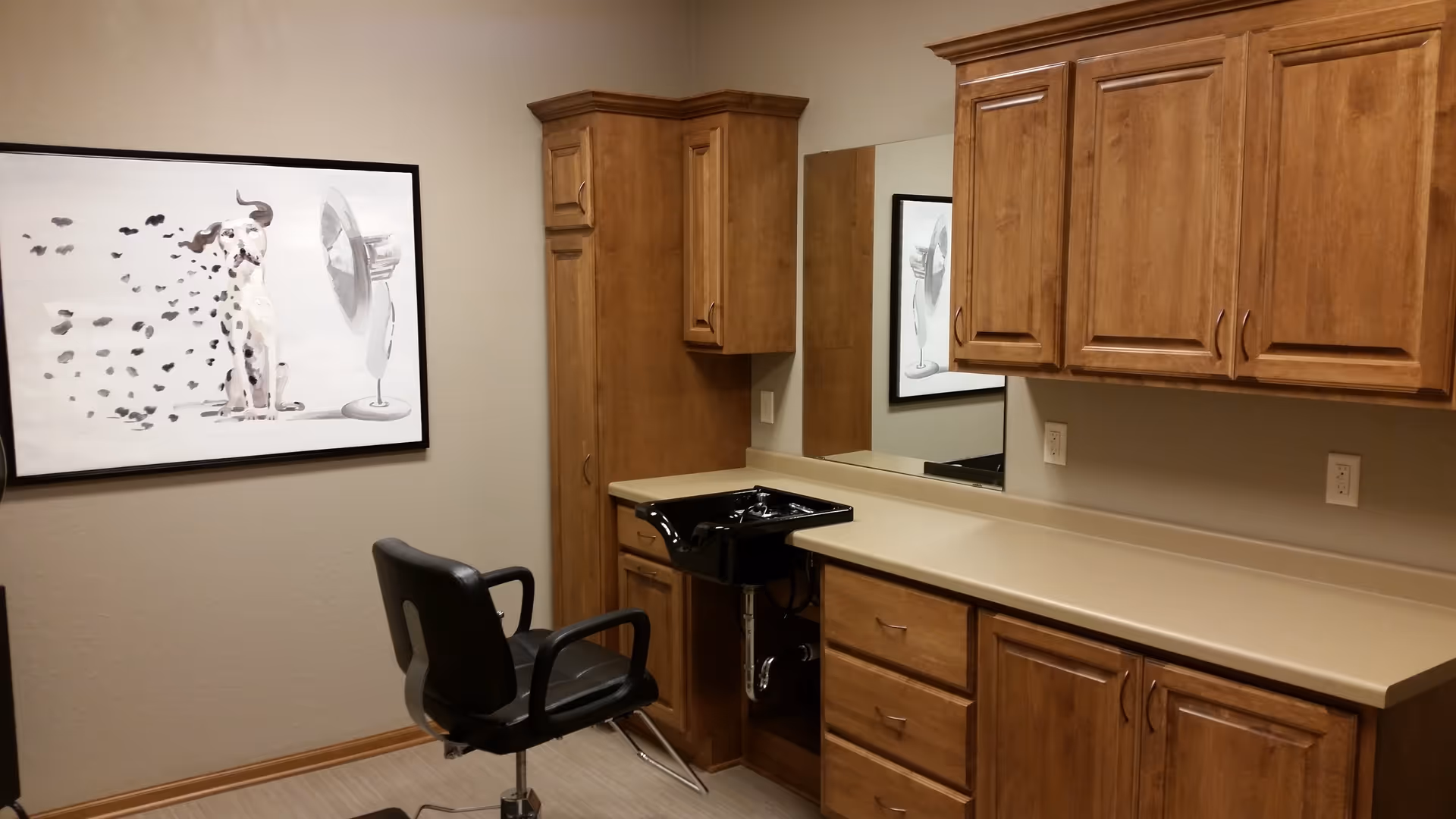 Interior of a hair salon room with wooden cabinets, a black salon chair, a black sink for washing hair, a large mirror on the wall, and a framed picture of a dog sitting in front of a fan.