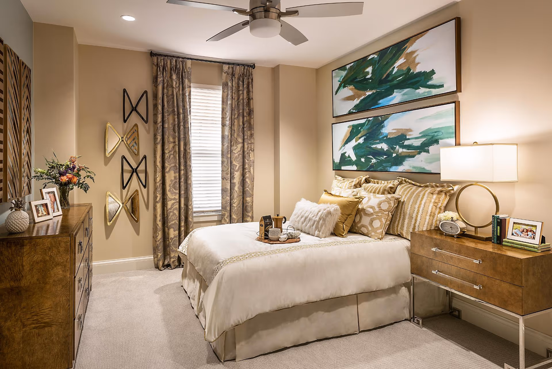 A cozy bedroom with a neatly made bed featuring multiple decorative pillows and a tray with a teapot and cups. The room has beige walls, a ceiling fan, patterned curtains covering a window, and two abstract green and white paintings above the bed. There is a wooden dresser with framed photos and a vase of flowers on the left, and a wooden nightstand with a lamp, clock, books, and a framed photo on the right.