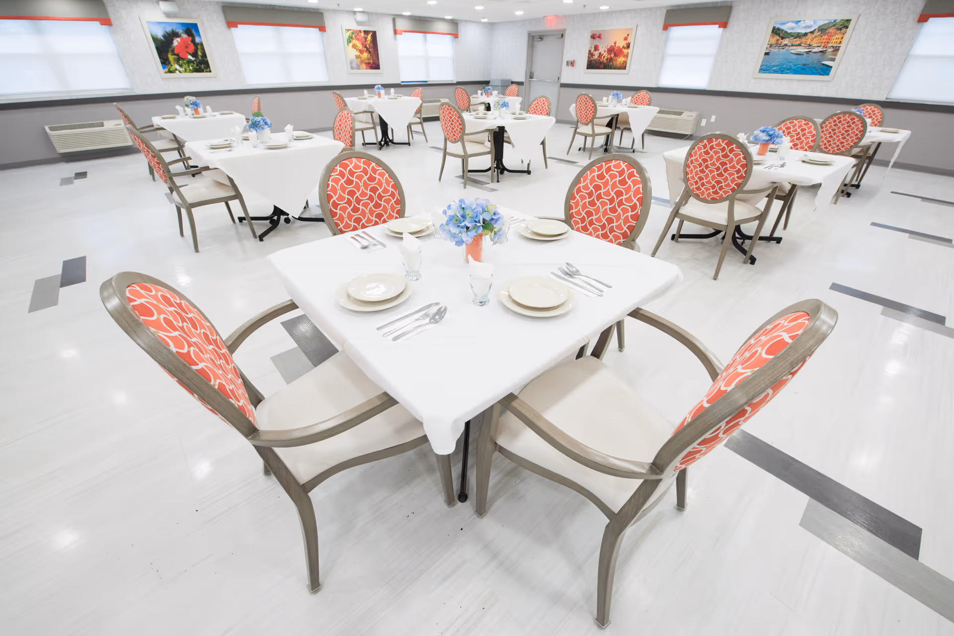 A bright and clean dining room with multiple tables covered in white tablecloths. Each table is set with plates, glasses, and silverware, and has a small floral centerpiece. The chairs have a wooden frame with red patterned upholstery. The room has large windows with blinds and colorful framed artwork on the walls.