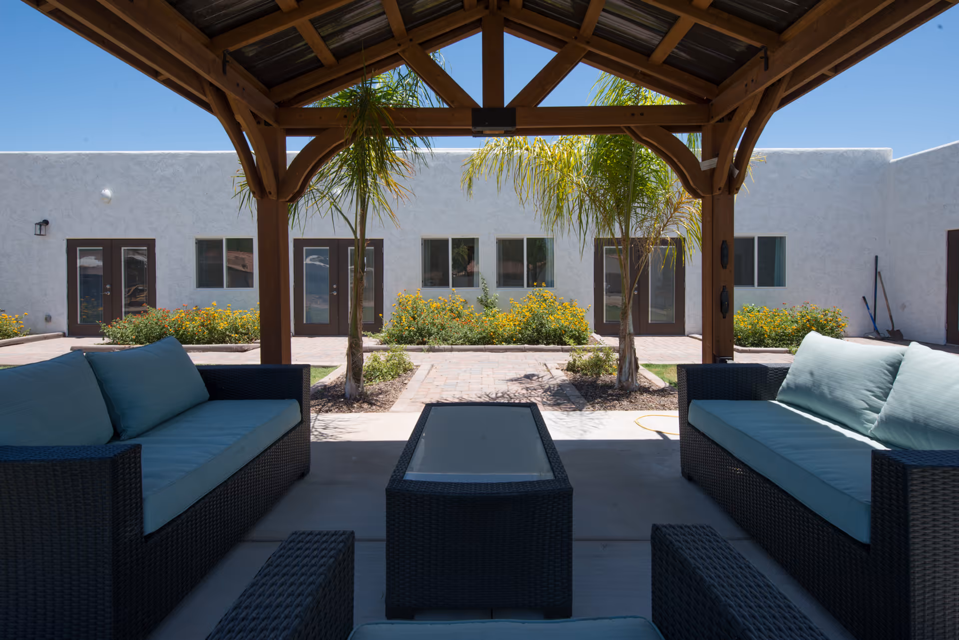 Covered outdoor patio with wicker sofas and a central glass-top table overlooking a courtyard and white stucco building.