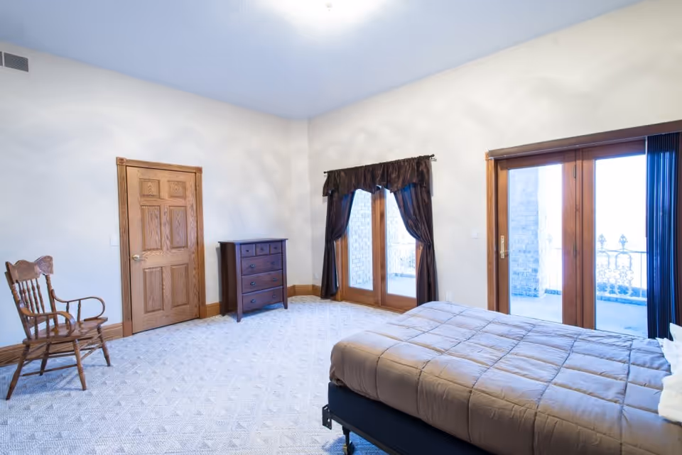 A bright bedroom with a bed covered in a brown comforter, a wooden rocking chair, a wooden dresser, and two sets of glass doors with dark curtains leading to an outdoor balcony.