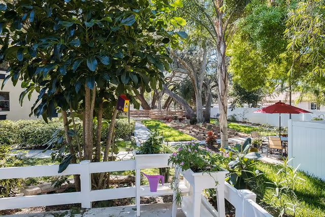 A sunny outdoor garden area with lush green trees, plants, and flowers. There is a white wooden fence in the foreground with a purple watering can placed on it. In the background, there is a patio area with a table, chairs, and a red umbrella, surrounded by more greenery and a white fence.