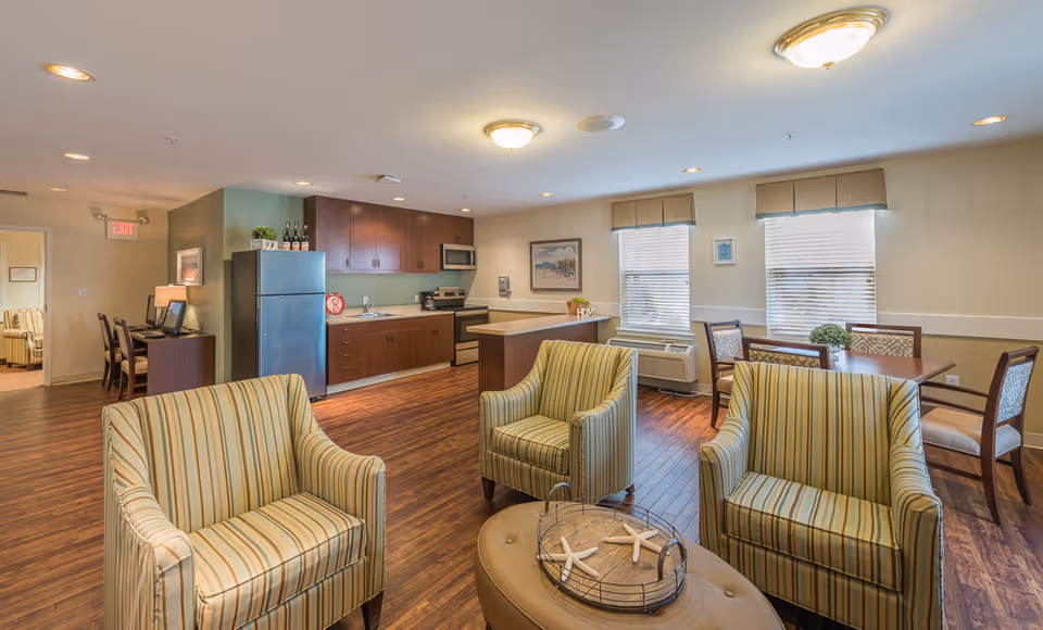 A bright and spacious common area in a senior living facility featuring a small kitchen with wooden cabinets, a refrigerator, microwave, and stove. In the foreground, there are three striped armchairs arranged around a round ottoman with decorative starfish. In the background, there is a dining table with four chairs near two windows with blinds and valances. The room has wooden flooring and soft lighting from ceiling fixtures.