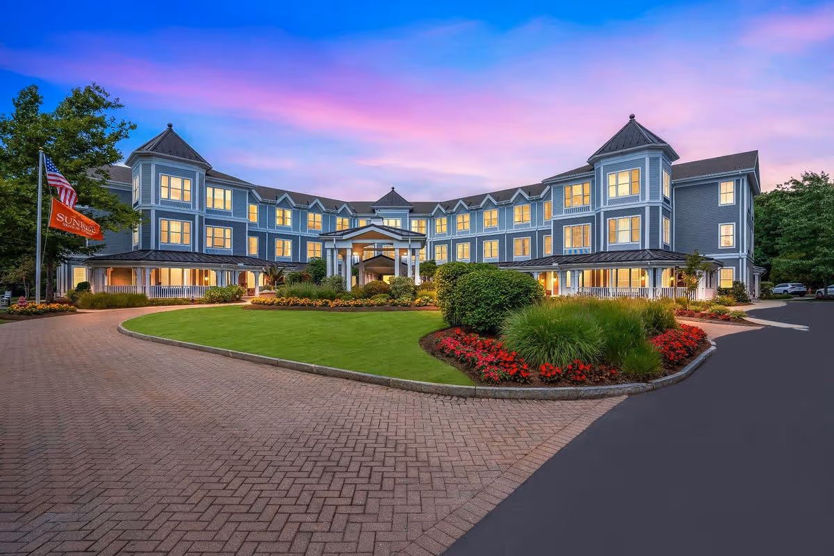 Exterior view of a large, three-story senior living facility building at sunset with a well-maintained lawn, flower beds, and a circular driveway. The building has multiple windows lit from inside and features two towers on each end. An American flag and a Sunrise flag are visible on the left side near the entrance.