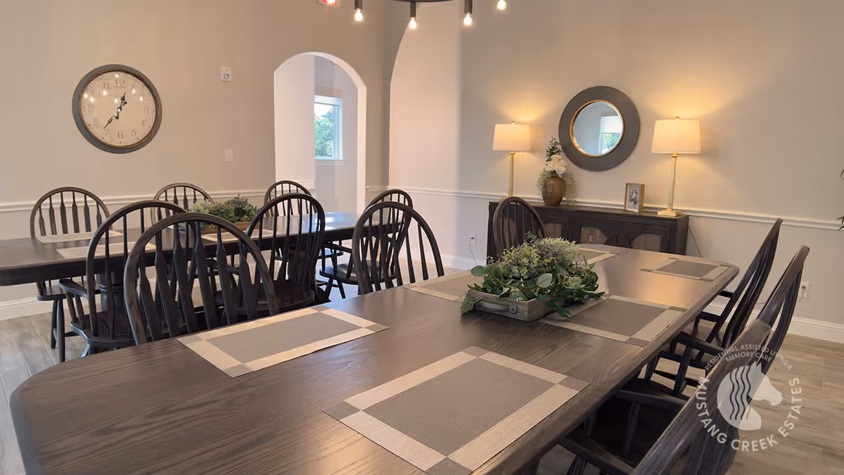 A dining room with two dark wooden tables arranged with beige placemats and a centerpiece of green foliage. The room features wooden chairs with vertical slats, a large round wall clock, a sideboard with two lamps, a round mirror, and a small framed picture. The walls are painted light beige, and there is an arched doorway leading to another room.