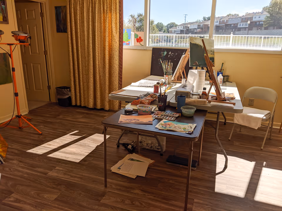 A bright art room with two tables covered in painting supplies including brushes, paint palettes, paper towels, and artwork. There is a large window letting in natural light and a folding chair with a towel draped over it. The room has wood flooring and a curtain covering a doorway.