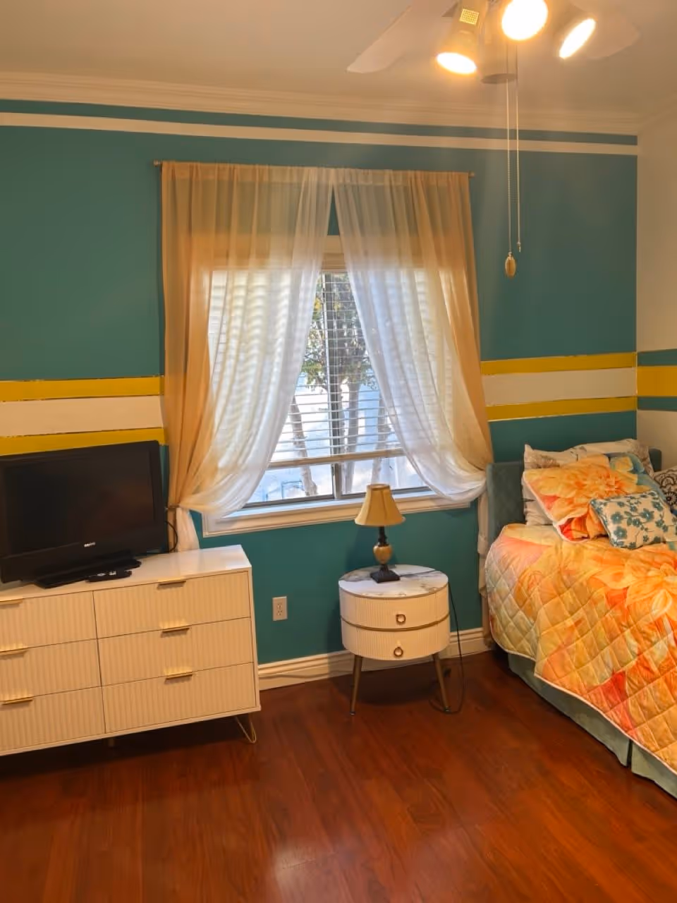 A bedroom with teal walls featuring yellow and white horizontal stripes. There is a window with sheer white curtains letting in natural light. Below the window is a small round white nightstand with a lamp on it. To the left is a white dresser with a small flat-screen TV on top. On the right side, there is a bed with a colorful floral quilt and multiple pillows. The floor is wooden, and a ceiling fan with lights is visible above.