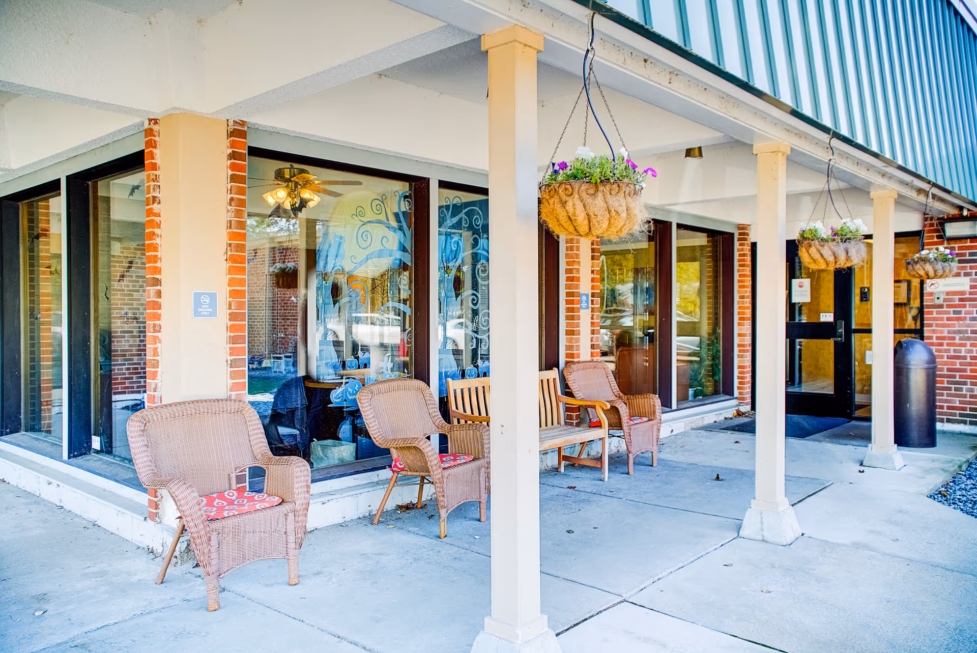Covered building entrance with wicker chairs, hanging flower baskets, and large windows reflecting the exterior.