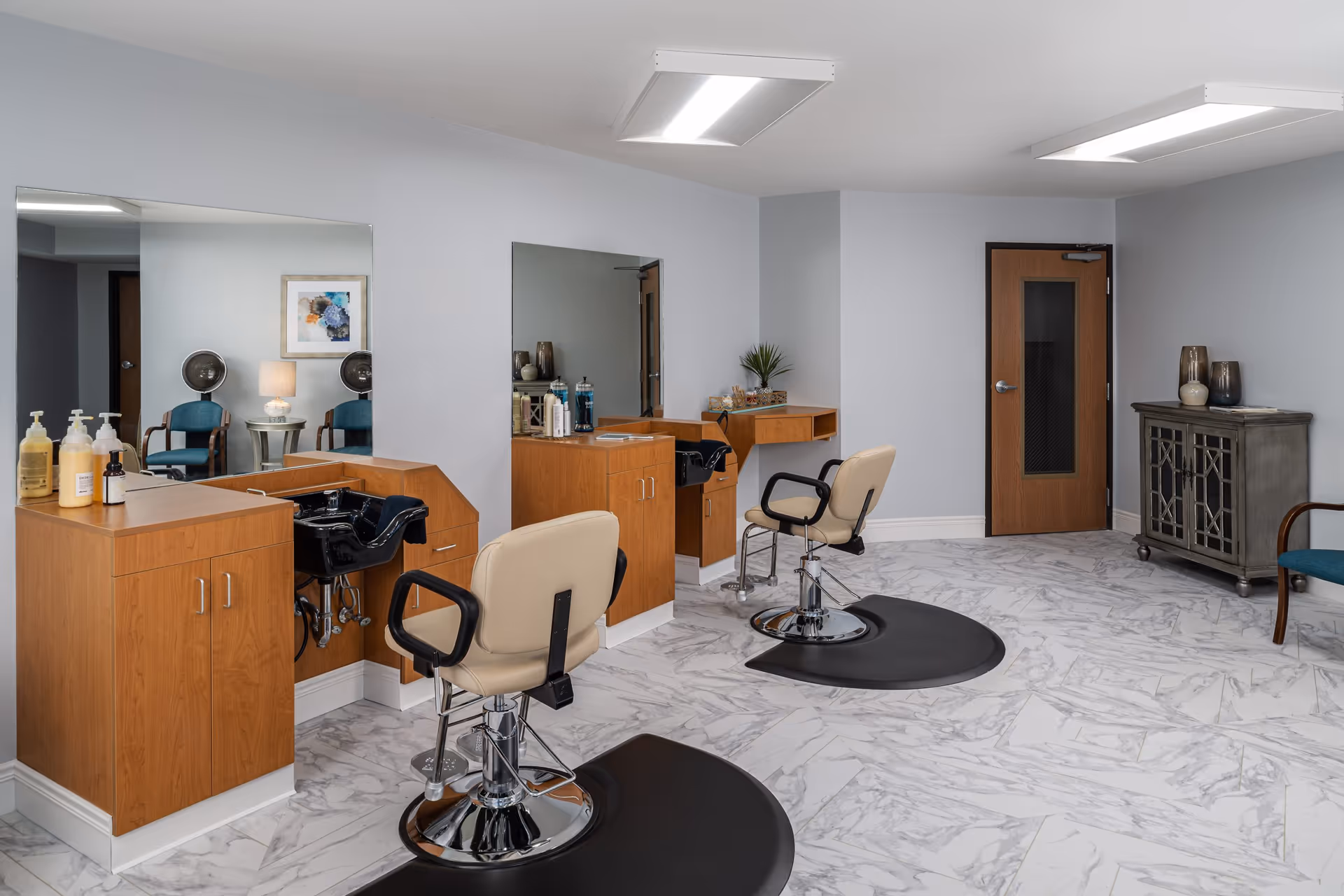 Interior view of a senior living facility's hair salon area featuring two beige salon chairs with black armrests in front of wooden cabinets with sinks and large mirrors. The floor has a white and gray marble pattern, and the walls are painted light gray. There is a wooden door, a gray cabinet with decorative vases, and a blue chair in the background.