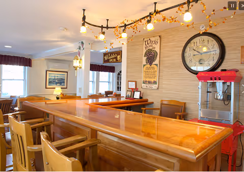 Interior view of a cozy assisted living facility bar area with a polished wooden counter and several wooden bar stools. The wall behind the bar features a large clock, a vintage-style wine bar sign, and a popcorn machine. Warm lighting fixtures hang from the ceiling, decorated with autumn leaves. In the background, there are windows with curtains and framed artwork on the walls.