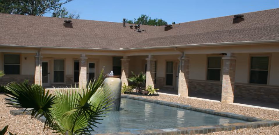 Outdoor courtyard area of a senior care facility with a rectangular water feature and a large decorative urn in the center. The courtyard is surrounded by a building with multiple doors and windows, beige walls, and stone pillars. There are small palm plants and gravel landscaping around the water feature under a clear blue sky.