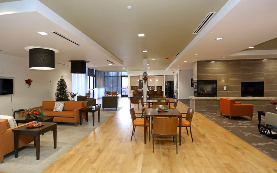 Spacious senior living facility common area with wooden flooring, multiple seating arrangements including orange sofas and wooden chairs around tables, a decorated Christmas tree, modern lighting fixtures, and a fireplace with a TV mounted above it.