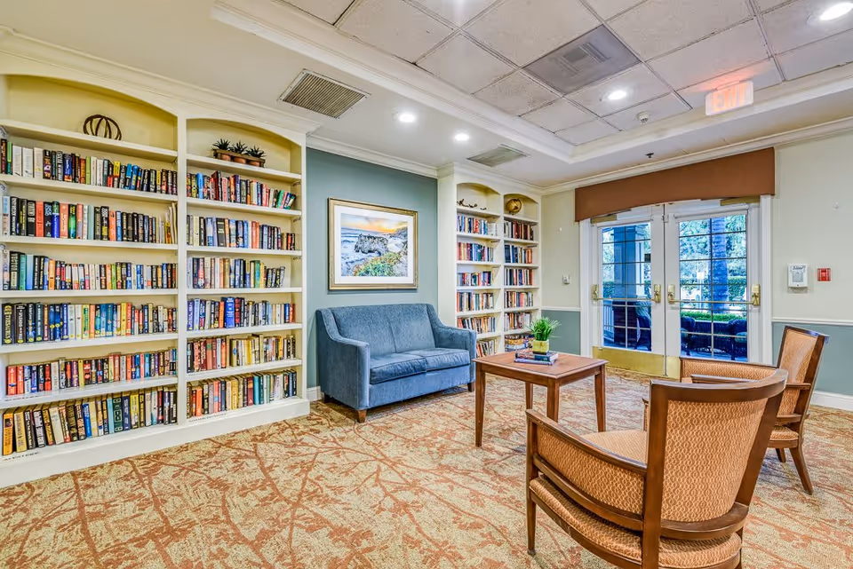 A cozy reading room with built-in bookshelves filled with books, a blue loveseat, two wooden armchairs with patterned cushions, and a wooden coffee table with a small plant and books on it. The room has a patterned carpet, a framed picture on the wall, and glass double doors leading outside.