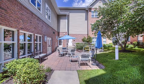 Sunny courtyard patio with tables, umbrellas, chairs, and landscaping next to a brick-faced building.