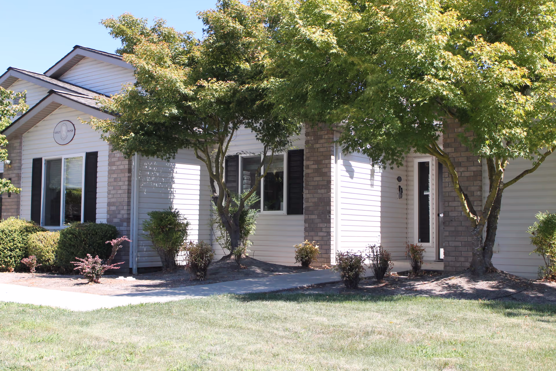 Exterior view of a single-story building with white siding and brick accents, surrounded by green trees and shrubs, under a clear blue sky.
