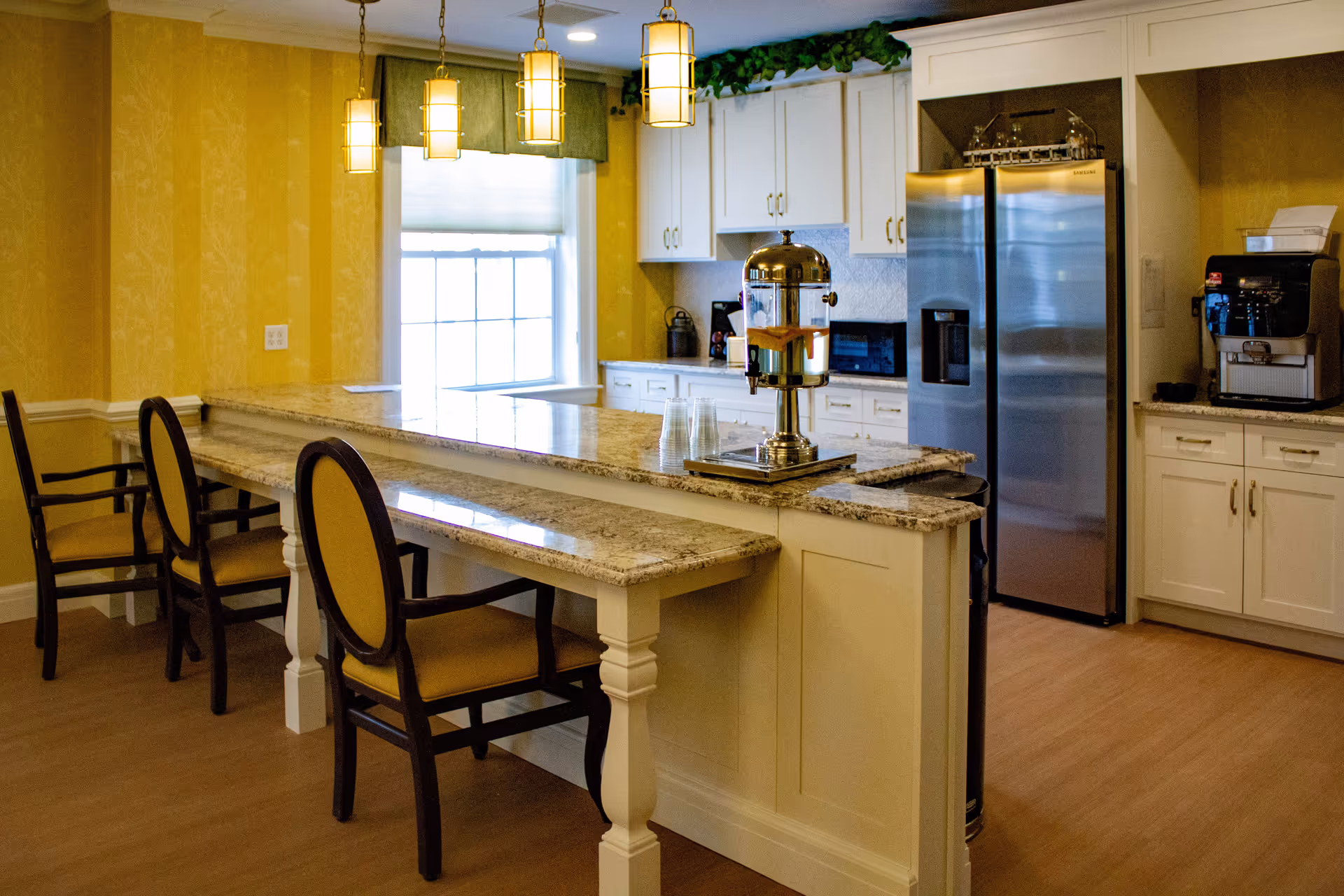 A bright kitchen area with yellow walls and white cabinetry. There is a large granite countertop island with three chairs, a beverage dispenser with cups on the counter, a stainless steel refrigerator, a coffee machine, and a window with a green valance letting in natural light.