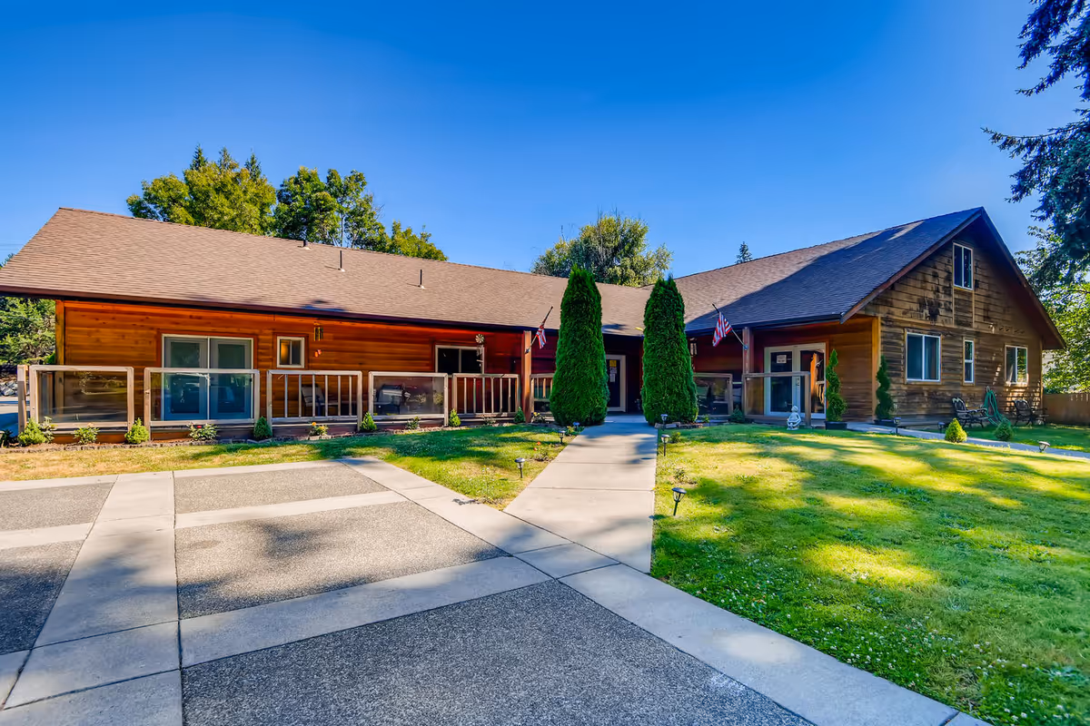 Exterior view of a single-story wooden building with a brown roof, surrounded by green grass and trees under a clear blue sky. The building has a paved walkway leading to the entrance, flanked by two tall evergreen shrubs and small American flags near the doors.