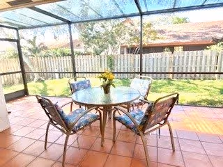 A screened-in patio area with a round glass table and four cushioned chairs. A vase with yellow flowers is placed on the table. The patio has terracotta tile flooring and overlooks a fenced backyard with green grass and trees.