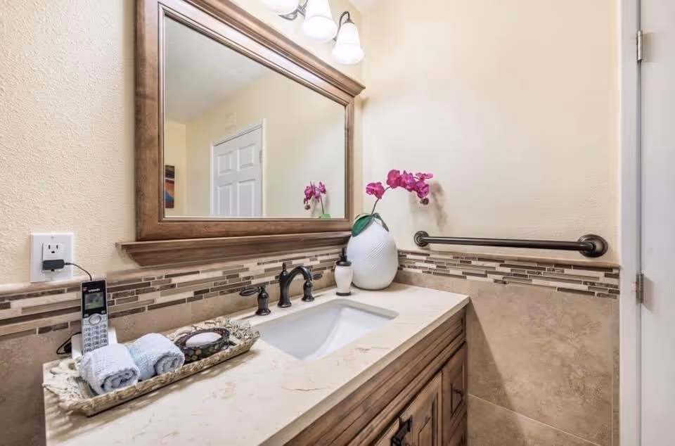A bathroom vanity with a large wooden framed mirror above it. The countertop has a white sink with black faucet fixtures, a white vase with pink flowers, a soap dispenser, and a tray holding two rolled towels and a small dish. There is a handrail on the wall next to the sink and a closed white door to the right.