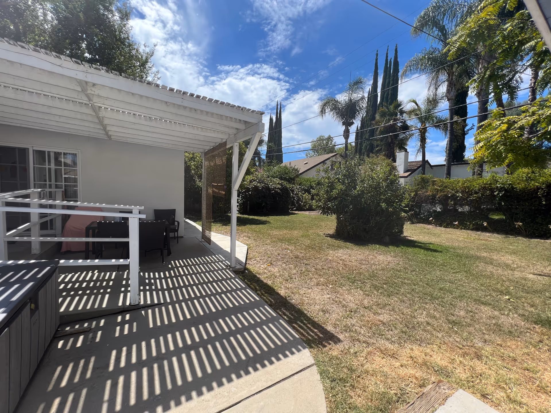 A sunny outdoor patio area with a white pergola casting striped shadows on the concrete floor. There is a black table with chairs under the pergola, next to a white building with sliding glass doors. The patio opens to a grassy yard with bushes, tall palm trees, and other greenery under a partly cloudy blue sky.