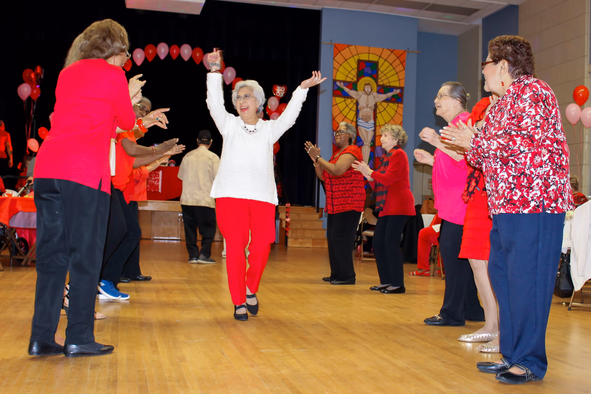 A group of elderly women standing in two lines facing each other in a large room, clapping and cheering as a woman in a white sweater and red pants joyfully walks down the middle with her arms raised. The room has wooden floors, a stage with red and pink balloons, and a large stained glass artwork of Jesus on the cross on the wall.