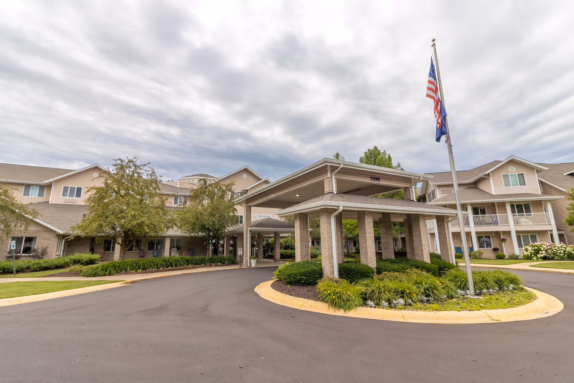 Exterior view of StoryPoint Wyoming senior living facility showing the main entrance with a covered drop-off area, landscaped roundabout with greenery, and an American flag on a flagpole. The building is two stories with beige siding and multiple windows under a cloudy sky.