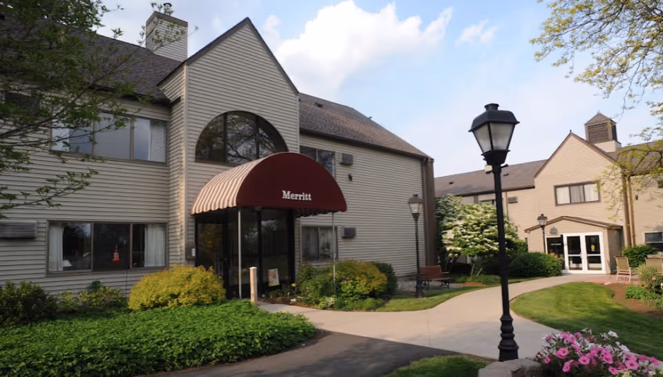 Exterior view of a senior living facility building with beige siding, a maroon awning labeled 'Merritt' over the entrance, surrounded by green bushes, trees, a walkway, and street lamps under a partly cloudy sky.