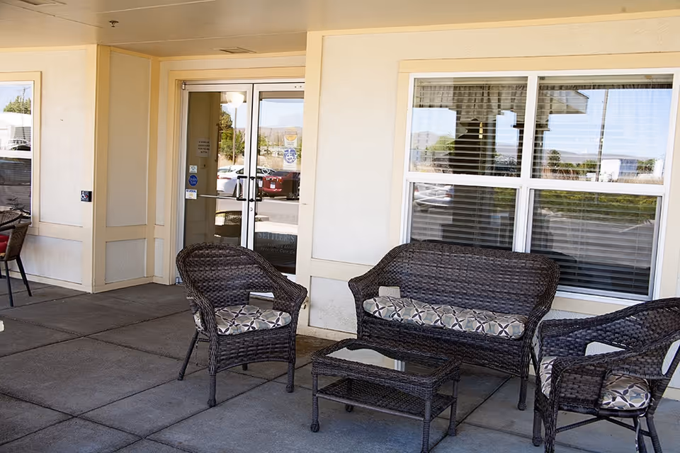 Covered entrance patio with wicker loveseat, chairs, and a small glass-top table in front of glass doors and windows.