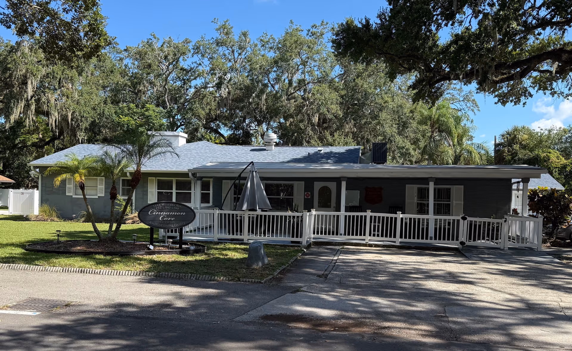 Single-story building with a gray exterior and a white railing ramp leading to the entrance. There are palm trees and other greenery around the building, and a sign in front reads 'Cinnamon Cove Assisted Living Facility'. The sky is clear and blue with some large trees in the background.