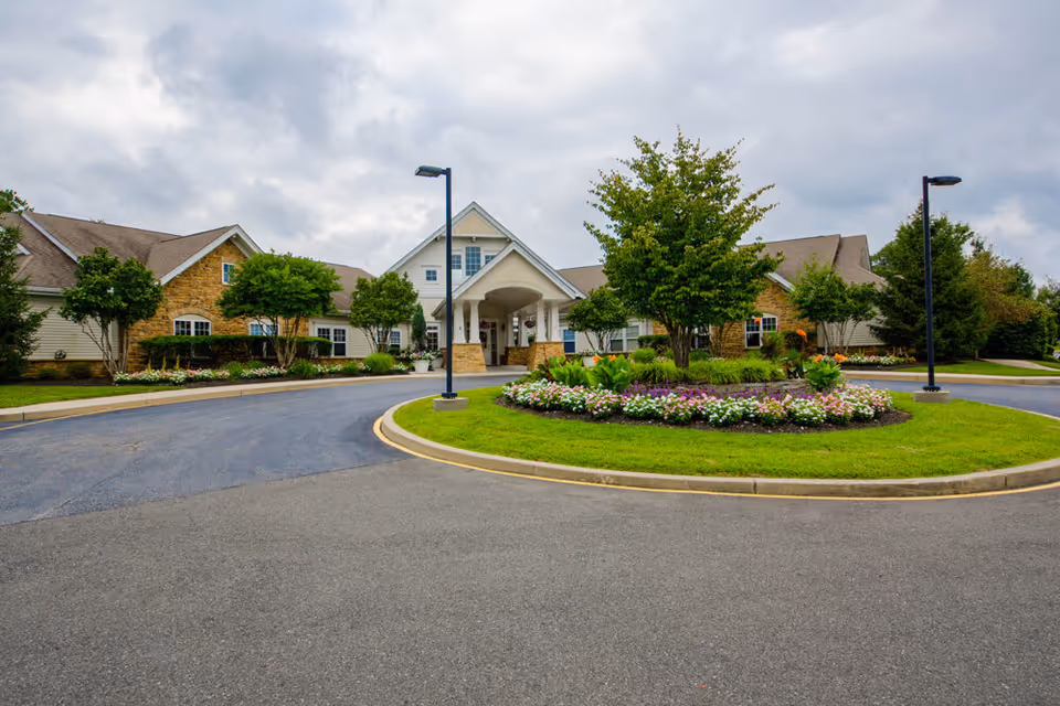 Front exterior view of Somerford House And Place Newark, featuring a circular driveway with a landscaped island of flowers and greenery, flanked by street lamps, and a building with a peaked roof and stone and siding facade under a cloudy sky.