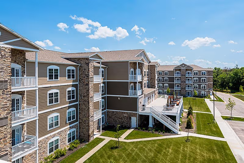 Exterior view of a multi-story senior living facility with beige and stone facade, balconies, a patio area with seating, well-maintained green lawns, and a clear blue sky with scattered clouds.