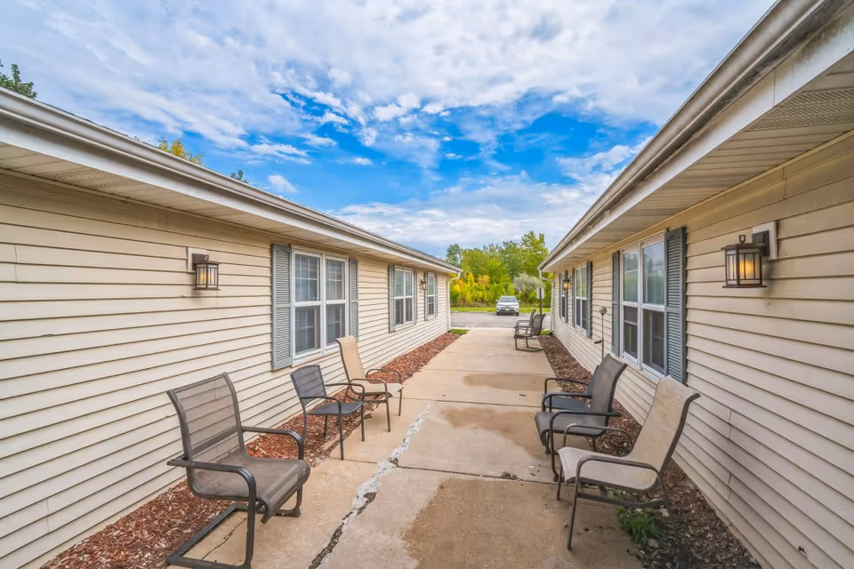 Outdoor walkway between two beige single-story buildings with windows and outdoor wall lights. Several metal chairs with mesh seats and backs are placed along the walkway. The sky is partly cloudy with blue patches visible.