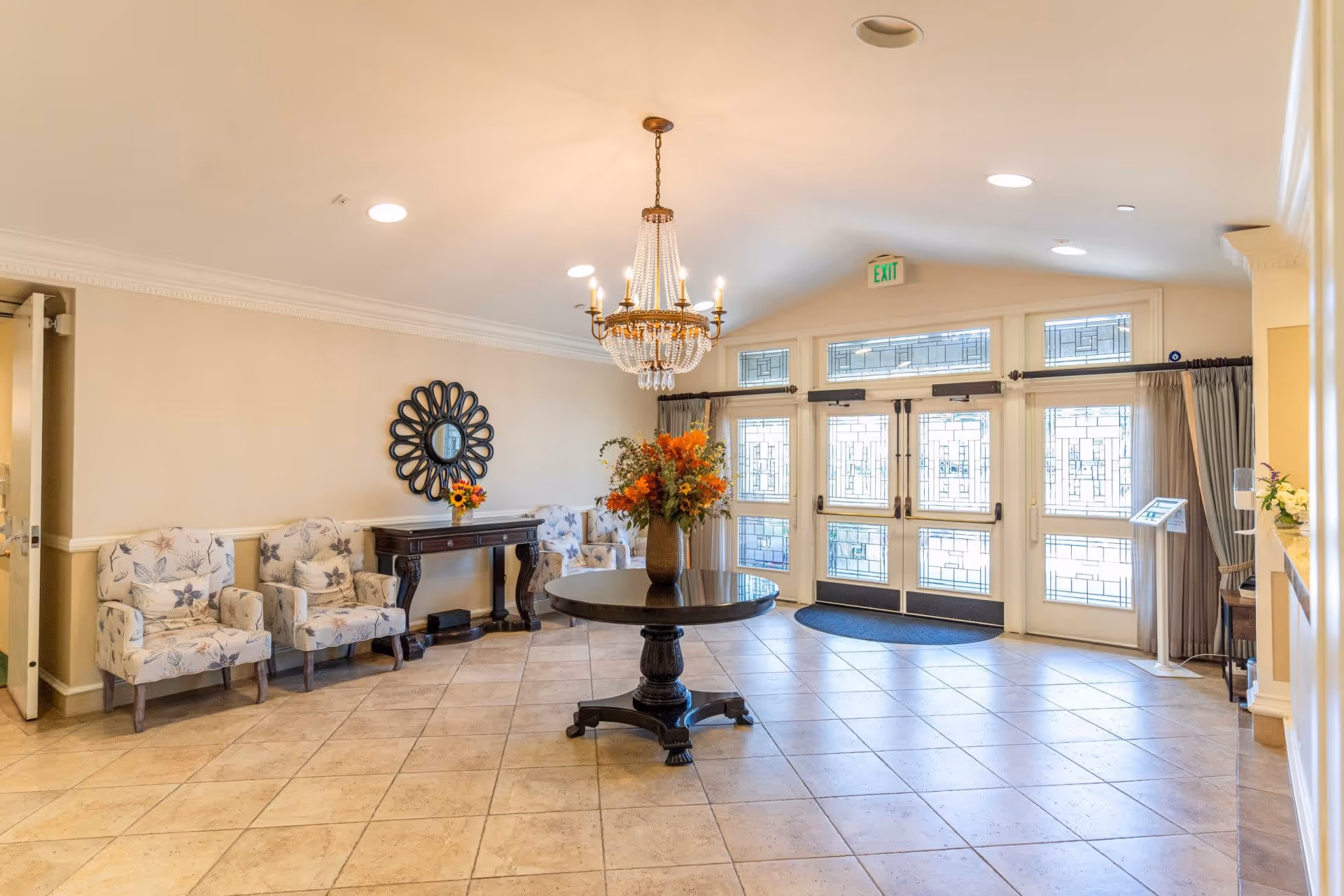Bright and spacious lobby area with a round wooden table in the center holding a large floral arrangement. There are four patterned armchairs along the left wall, a decorative mirror above a dark wooden console table, and large glass double doors with intricate designs letting in natural light. A chandelier hangs from the ceiling, and the floor is tiled.