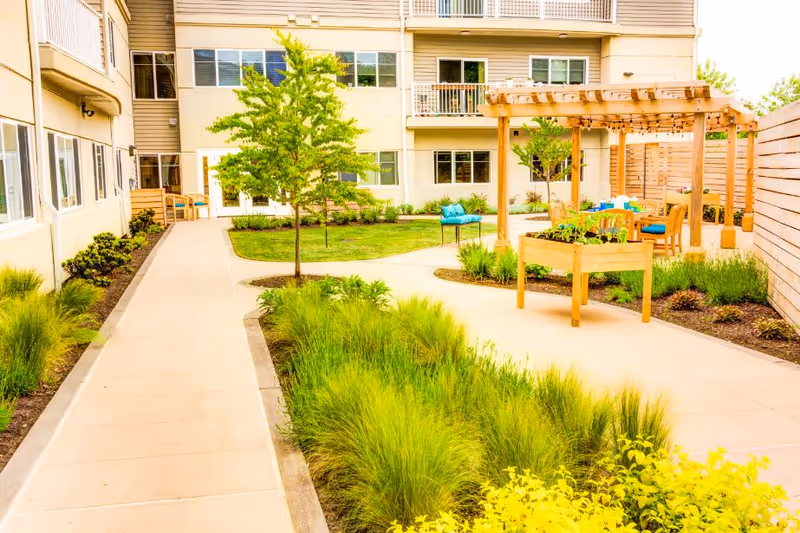 Outdoor courtyard area at Harbour Pointe Senior Living featuring a paved walkway, green grass, small trees, and a wooden pergola with string lights. There are wooden raised garden beds and a seating area with chairs and a table under the pergola. The courtyard is surrounded by a multi-story building with windows and balconies.