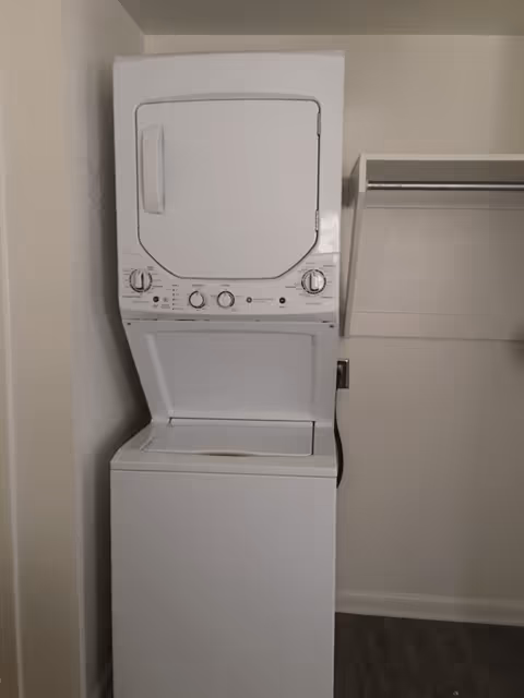 A stacked white washer and dryer unit in a small laundry area with a shelf and hanging rod on the right side.