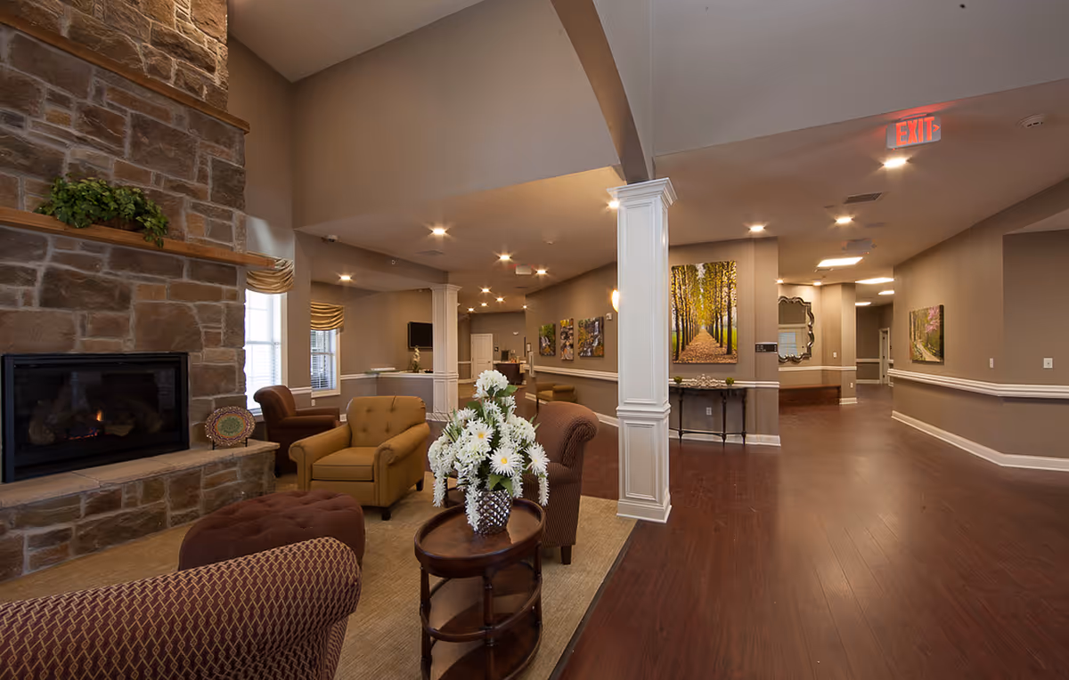 Spacious senior living lounge with a stone fireplace, upholstered chairs around a small table with flowers, and a wide hallway leading past artwork.