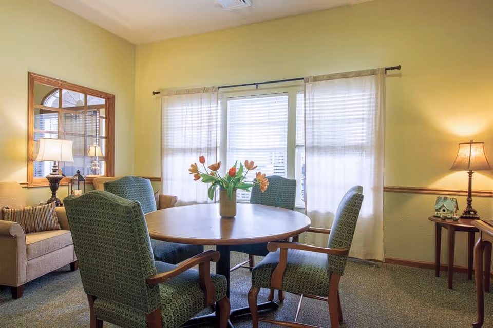 A cozy sitting area with a round wooden table surrounded by four green upholstered armchairs. A vase with orange tulips is placed on the table. The room has light yellow walls, a window with sheer white curtains, and two table lamps on either side providing warm lighting. There is a beige armchair with a striped pillow and a wooden side table with a small decorative house.
