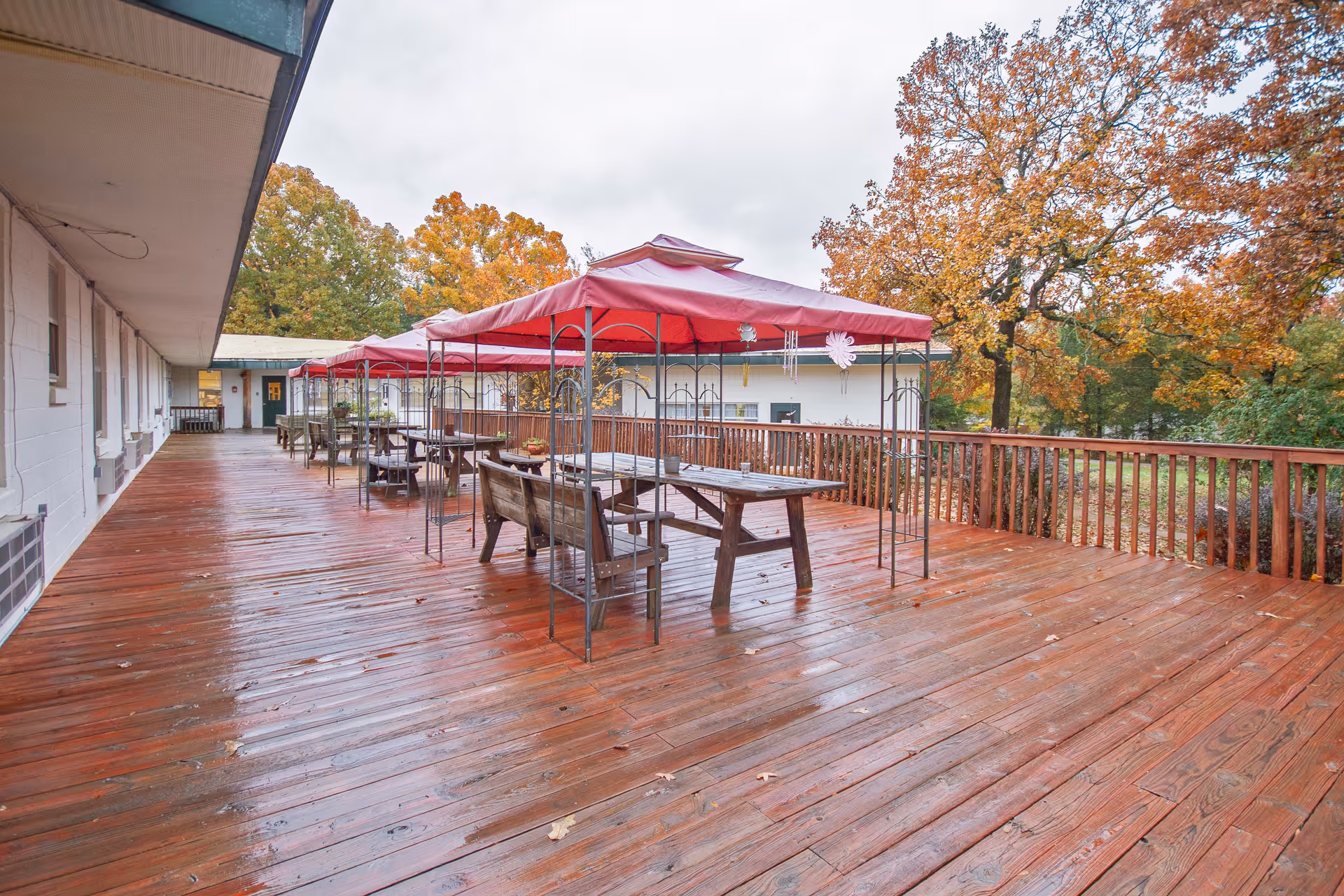 A large wooden outdoor deck with picnic tables under red canopies and autumn trees beyond a railing.