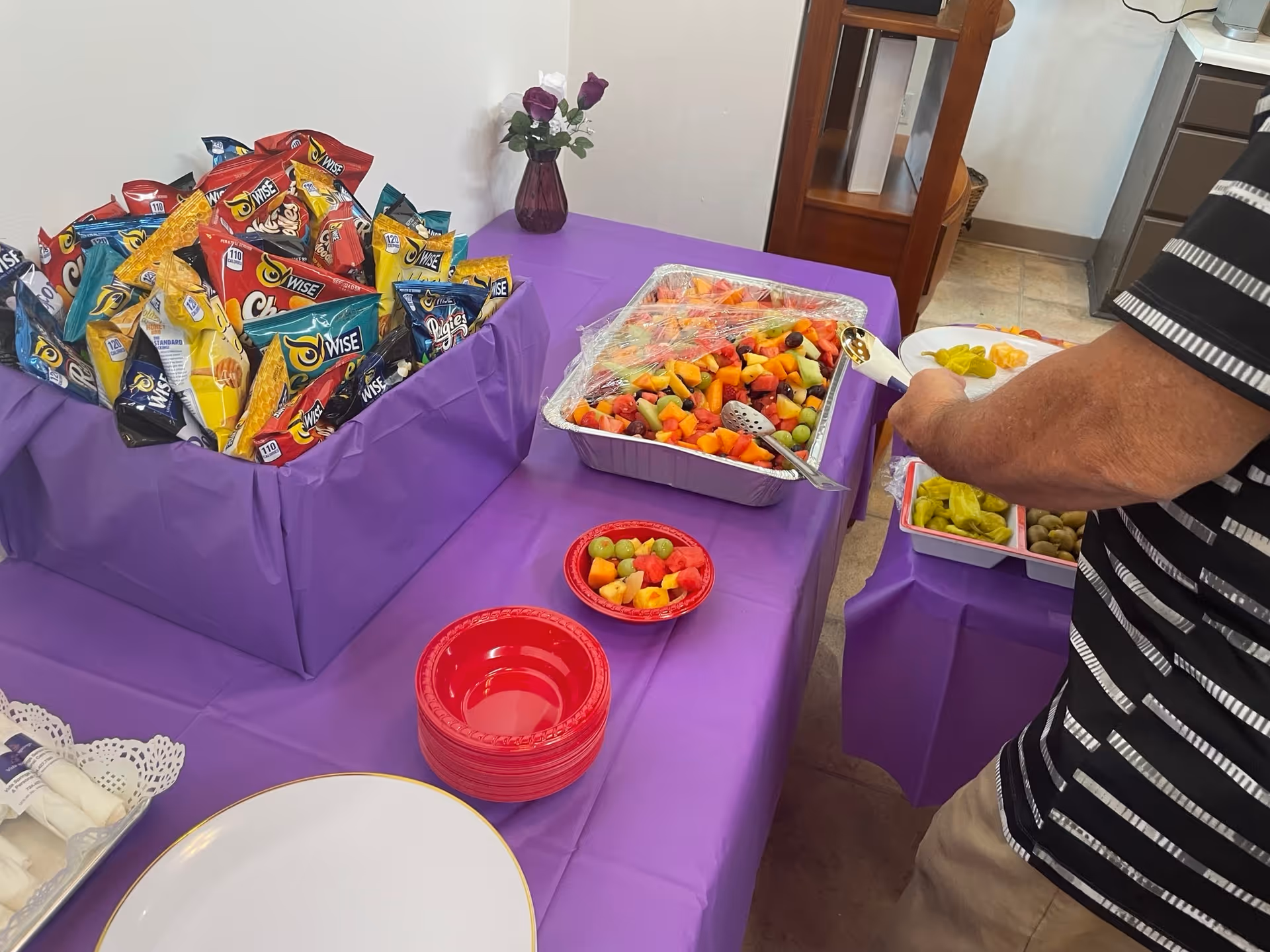 A purple-covered buffet table with bags of chips, a tray of mixed fruit, red bowls, and a person serving themselves.