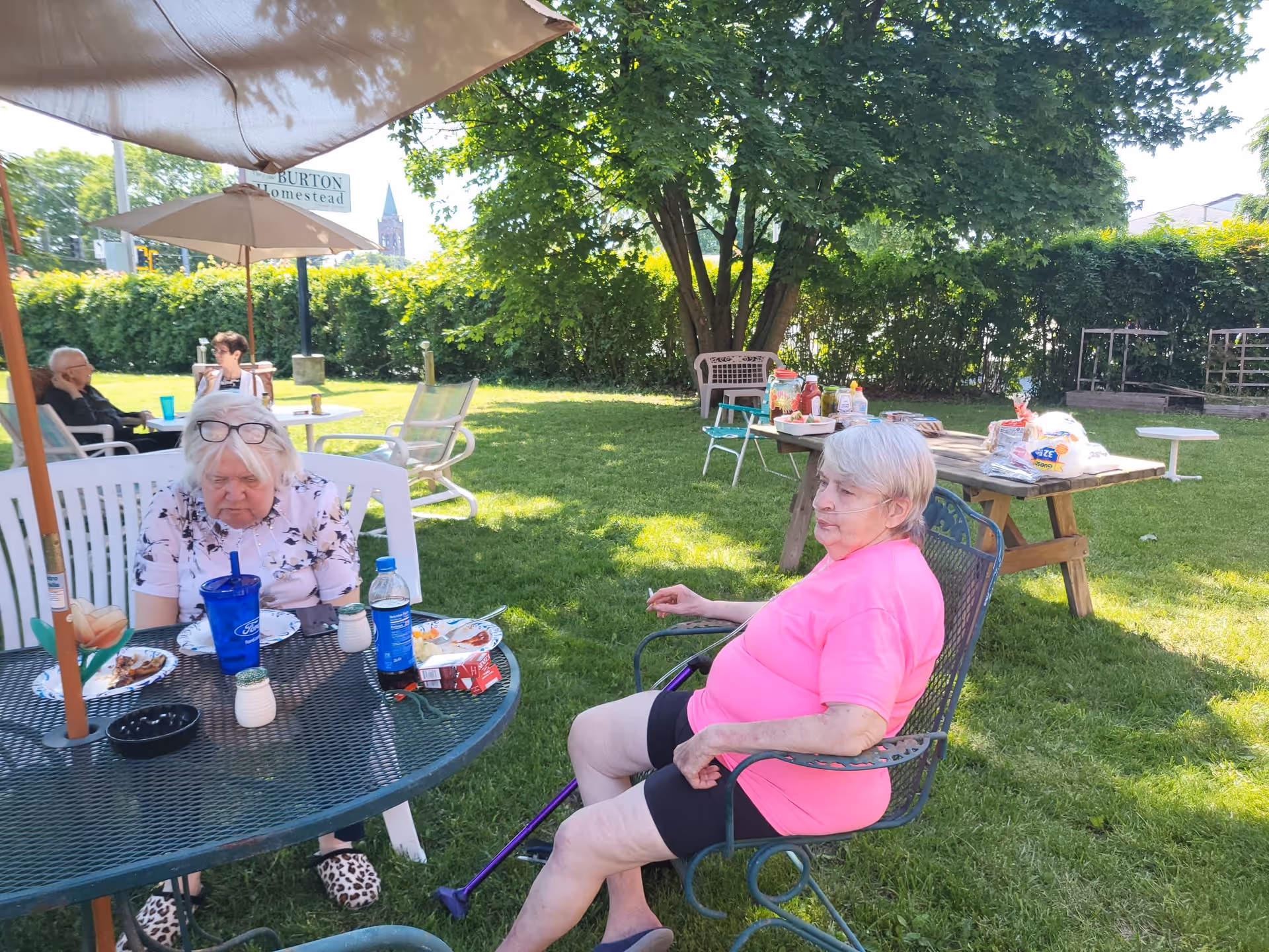 Two elderly women sitting at a round outdoor table with food and drinks, under an umbrella in a grassy yard. One woman is wearing a pink shirt and black shorts, sitting on a metal chair with a cane beside her. The other woman is wearing a floral shirt and glasses, sitting on a white plastic chair. In the background, there are more people sitting at another table, a picnic table with condiments and food items, trees, and a sign that reads 'Burton Homestead'.