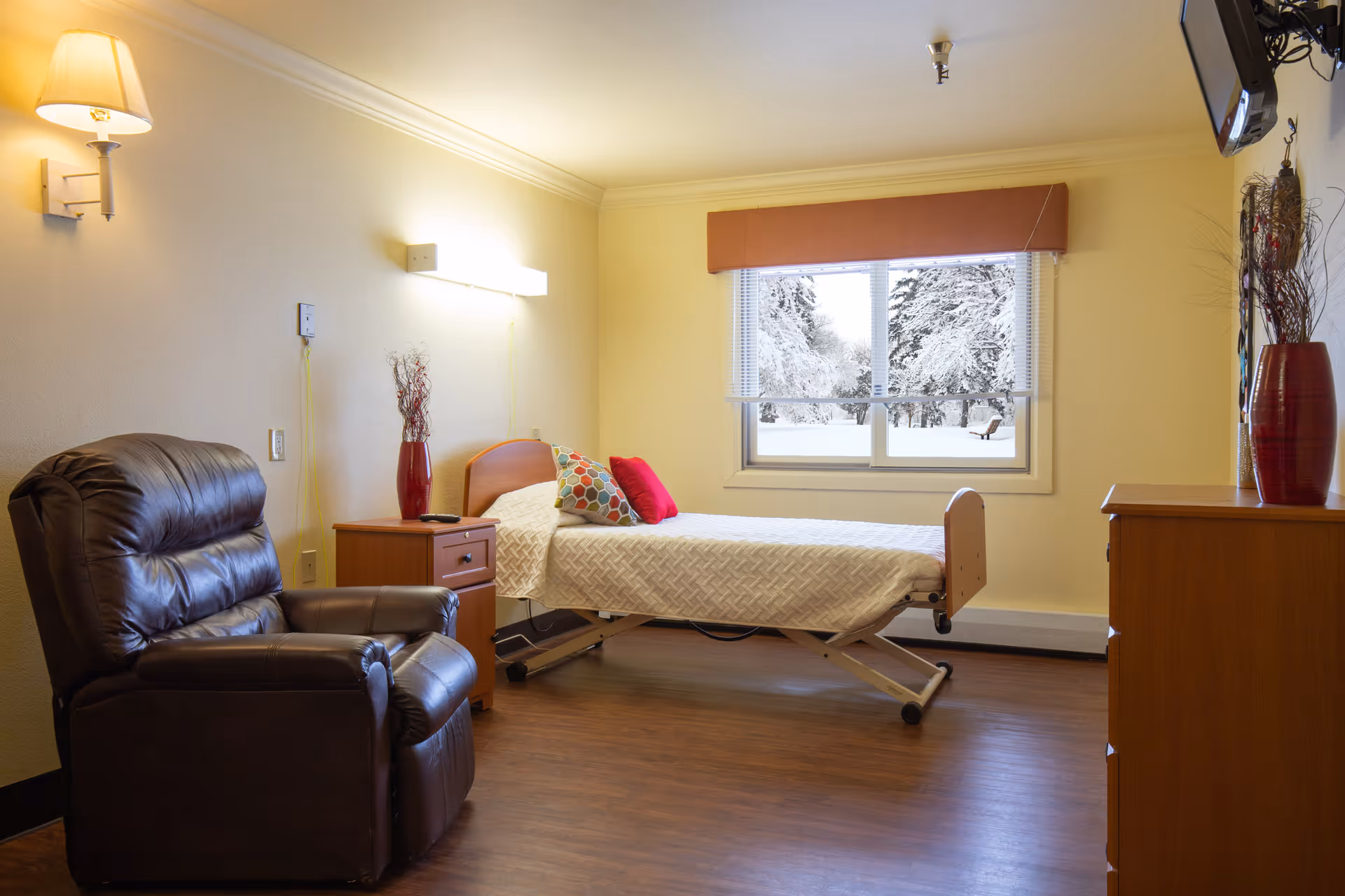 A cozy bedroom in a senior living facility featuring a single adjustable bed with patterned bedding and colorful pillows. Next to the bed is a wooden nightstand with a red vase holding decorative branches. A large, comfortable dark brown leather recliner is positioned near the nightstand. The room has wooden flooring, soft yellow walls, a wall-mounted lamp, and a window with a view of snow-covered trees outside. A wooden dresser with another red vase and decorative branches is on the right side, and a TV is mounted on the wall above it.