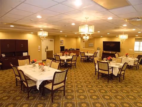 A dining room in an assisted living facility with several tables covered with white tablecloths, each surrounded by chairs. The room has patterned carpet flooring, warm lighting from ceiling fixtures, and a large flat-screen TV mounted on the wall. There are flower centerpieces on the tables and cabinets along one wall.