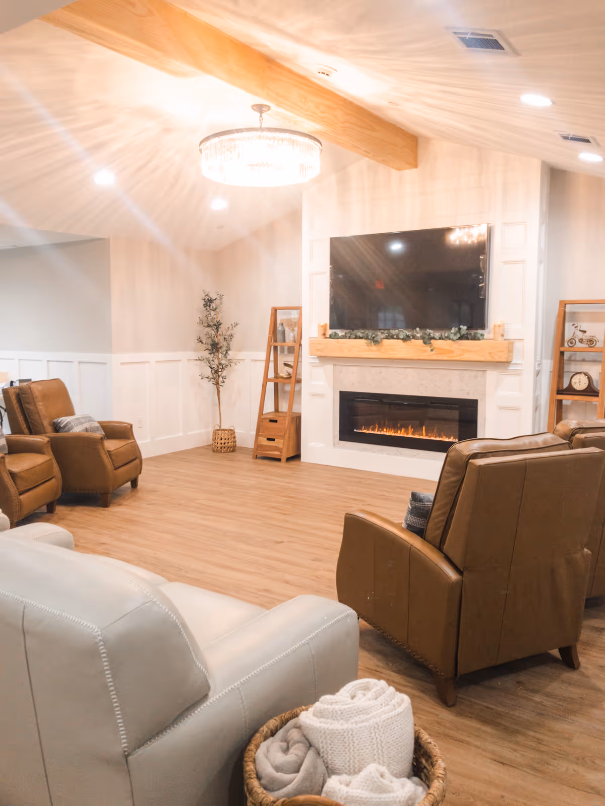 A cozy living room with a modern electric fireplace beneath a mounted flat-screen TV. The room features wooden flooring, a wooden beam on the ceiling, and a chandelier light fixture. There are several comfortable leather armchairs arranged around the fireplace, a basket with folded blankets in the foreground, and decorative shelving units with small plants and ornaments on either side of the fireplace.