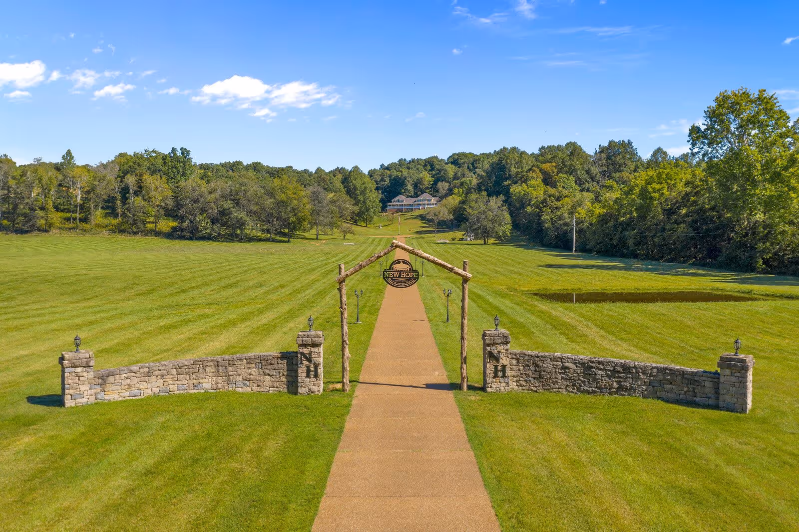 A long paved pathway flanked by green grass and trees leads to a large building in the distance. At the start of the pathway, there is a rustic wooden arch with a circular sign that reads 'New Hope Senior Living of Hendersonville.' Stone walls with lanterns are on either side of the pathway entrance. The sky is clear with a few clouds.