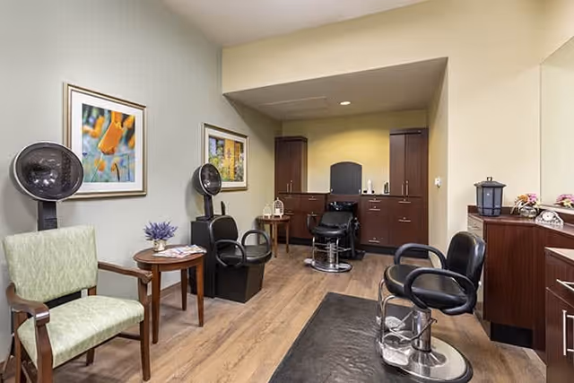 Interior of a salon room with three black salon chairs, two hair dryer stations, wooden cabinets, a small table with a flower vase, and framed floral artwork on the walls. The floor is wooden and the room has a warm, inviting atmosphere.